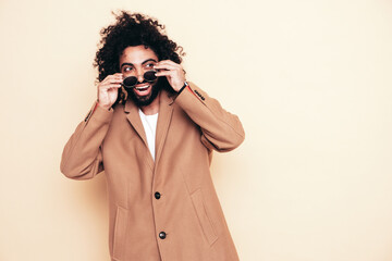 Handsome smiling hipster model.  Unshaven Arabian man dressed in brown coat clothes. Fashion male with long curly hairstyle isolated in studio. Cheerful and happy. In sunglasses