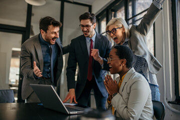 Team of people standing around a laptop in the bright office and celebrating their achievement.