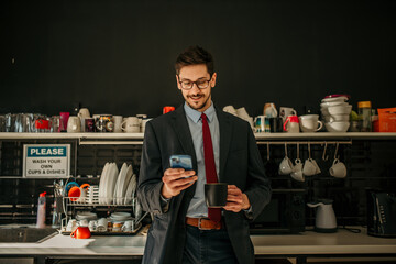 Businessman standing in the office kitchen and using a phone during a coffee break. - obrazy, fototapety, plakaty