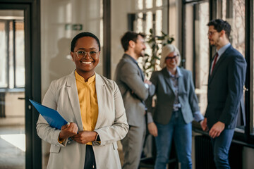 Portrait of a black businesswoman standing in the office, people in the background talking.