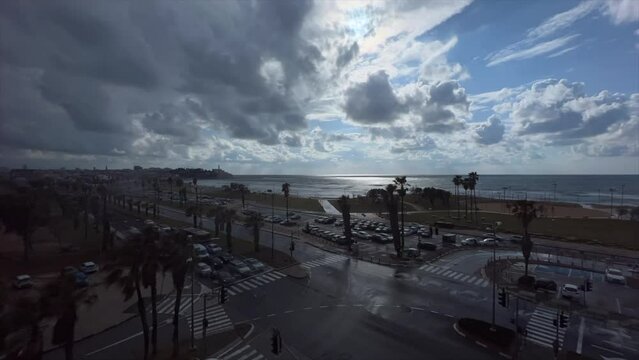 A time lapse view of the Tel Aviv coast, looking towards Jaffa. Storm clouds are rolling in from the Mediterranean, and traffic and pedestrians pass by.