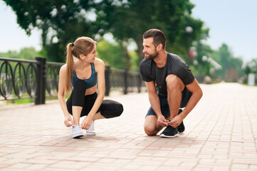 Image of young happy smiling couple, woman training with man or bearded coach trainer tying shoe laces, before morning jogging, cardio workout outdoors. Fitness, sport, healthy lifestyle concept.