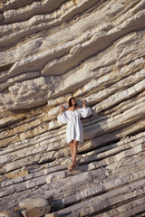 woman in white dress leaning on rock ledge, in the style of creased crinkled wrinkled, playful body manipulations, wide angle lens