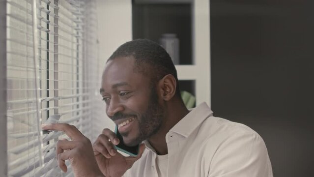 Chest up of handsome Black man talking on mobile phone and looking through window blinds while working in minimalist office