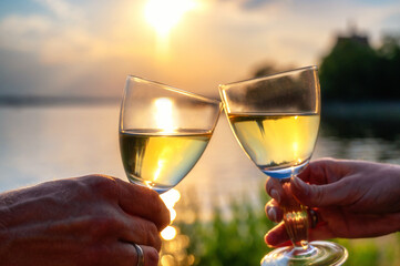 Ein Mann und eine Frau stoßen mit einem Glas Weißwein am Ufer von einem See an - obrazy, fototapety, plakaty