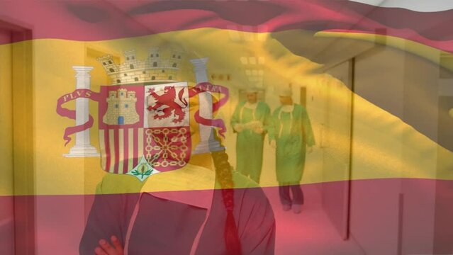 Animation of flag of spain, biracial female surgeon standing arms crossed in corridor of hospital