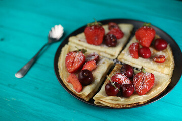 delicious fragrant pancakes folded in a triangle on a black plate and aquamarine wooden background and decorated with delicious ripe sweet strawberries, red fragrant cherries, coconut flakes and honey
