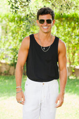 Happy handsome young male in white pants and sunglasses looking at camera confidently while standing against green plants in summer garden.