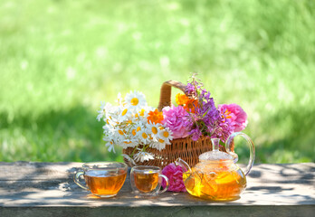 Wild flowers in basket, glass teapot and cups with herbal tea on table in garden, natural...
