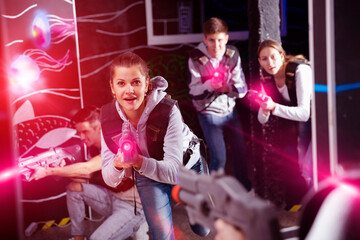 Excited girl aiming laser gun at other players during lasertag game in dark room..