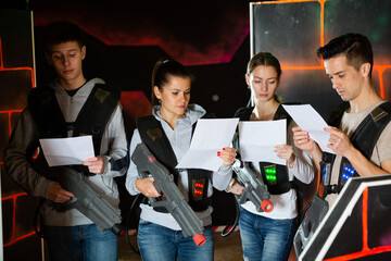 Group of young people wearing special vest and holding laser guns reading instruction before lasertag game