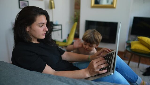 Child wanting mother attention sitting on sofa working from home. Parent multi tasking in front of laptop computer. Toddler baby bothering mom. Cute modern parenting lifestyle