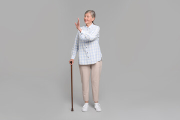 Senior woman with walking cane waving on gray background