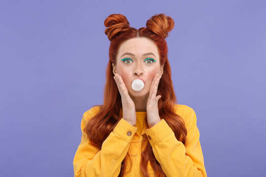 Portrait of surprised woman with bright makeup blowing bubble gum on violet background