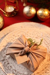 Furoshiki technique. Gift packed in beige fabric, blank card and festive decor on red table