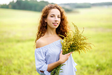 Red-haired girl with bouquets of yellow flowers in summer field. Young lady holding bouquet of wild flowers