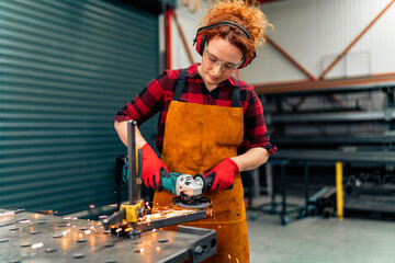 A young girl who is an apprentice in a metal workshop uses a grinder and works on a metal bar, she is wearing protective equipment
