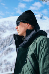 Young adventurous man in weather proof gear outfit, waterproof jacket, wool beanie hat and mountaineering sunglasses, look at mountain top, ready to summit. Ski touring and alpine skiing concept