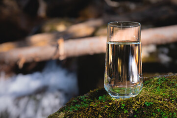 Purified fresh drinking water in a glass goblet from a mountain lake stands in the sunlight on a stone covered with moss, against the backdrop of a waterfall