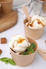 Scoops of ice cream with caramel sauce, mint leaves and candies on white tiled table, closeup