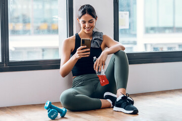 Sporty young woman using her mobile phone while holding protein shake after gym exercicis at home