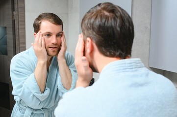 Handsome young man applying cream on his face in bathroom