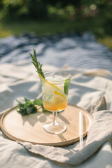 Old-fashioned glass with fresh mocktail garnished with lemon and rosemary twig on wooden tray at a picnic in nature.