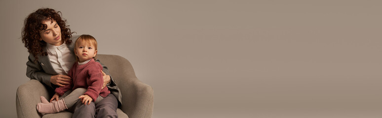 balancing work and life concept, curly woman in suit sitting in armchair with toddler daughter, grey background, multitasking, quality family time, mother and child, modern working parent, banner