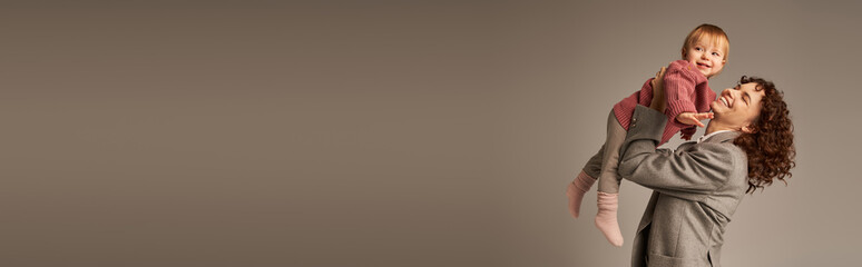 happy working mother, parenting and career, cheerful businesswoman lifting her toddler daughter on grey background, work life harmony concept, loving motherhood, quality time, joy, banner