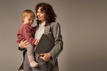 professional aspirations, modern parenting, work life harmony, cheerful working mother holding laptop and daughter on grey background, time management, self-confidence