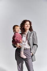 career and family, happy working mother holding toddler daughter in arms and standing with hand in pocket on grey background, work life balance, modern parenting, professional success, businesswoman