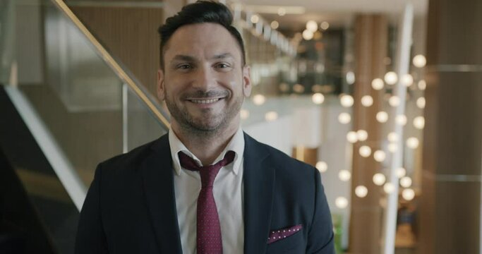 Potrait of joyful young businessman standing in modern hotel lobby and looking at camera smiling. People and positive emotions concept.