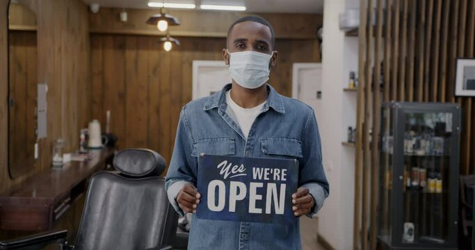 Portrait of barbershop owner wearing medical mask holding Yes we are open sign welcoming clients during covid-19 pandemic. Healthcare and business concept.