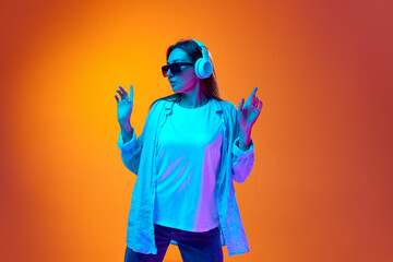 Portrait of young woman in casual clothes and sunglasses listening to music in headphones against gradient orange studio background in neon light