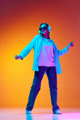 Young woman in casual clothes and sunglasses listening to music in headphones against gradient orange studio background in neon light