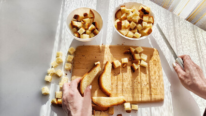 Homemade cooking and cutting of slices of diced dry white rye bread on cutting board and white plate. Hand of woman with Crackers for food, healthy eating on table in sunny day, top view