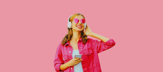 Portrait of stylish modern happy young woman in headphones with smartphone listening to music wearing jacket, sunglasses on pink background