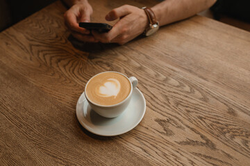 Cup of coffee on wooden table next to a man using cell phone. Coffee break in a city cafe.
