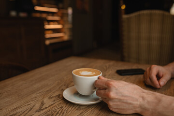Close-up of man holding a cup of cappuccino on wooden table in cafe. Copy space.