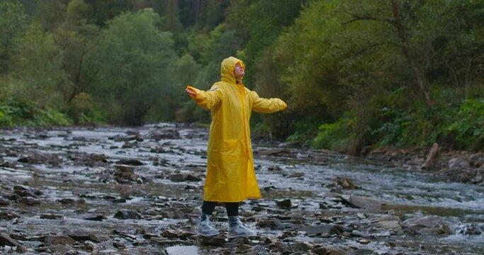 Happy young woman in yellow rain coat standing on mountain stream raising arms in slow motion. Wide shot portrait of confident relaxed Caucasian hiker outdoors. Cinema 4k ProRes HQ