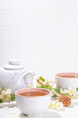 Organic herbal health drink. Jasmine flowers green tea, in white mugs, with teapot, on white table, with blooming jasmine flowers 