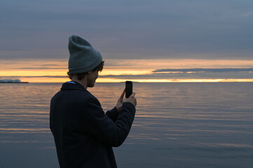 Man in coat and hat is taking picture on the phone of sunset above the sea or ocean. Iceland tourist