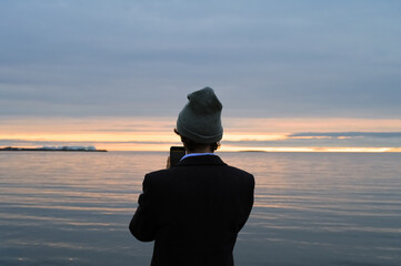 Man in coat and hat is taking picture on the phone of sunset above the sea or ocean. Iceland tourist