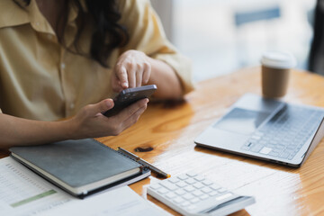 Fototapeta premium Asian businesswoman using mobile phone to search information and chat via application while sitting at work using laptop with paperwork making plan, investing, analyzing financial report.