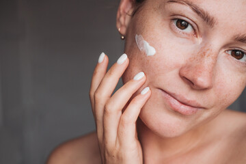 Young woman with cream on her cheek touching her face and looking at the camera, beauty routine 