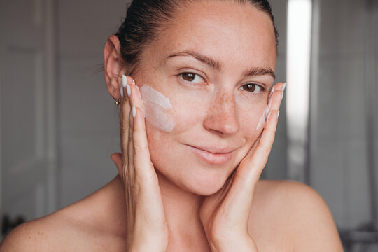 A young woman applying cream on her face with two hands, smiling and looking at the camera 