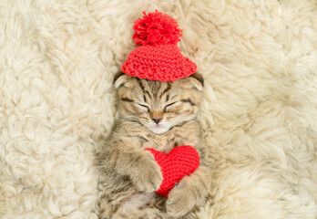 Cute fold tabby kitten wearing warm hat hugs red heart on the bed at home. Valentines day concept. Top down view