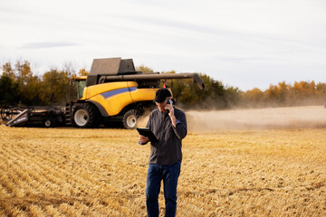 Farmer using a tablet to manage his harvest and talking on his cellphone with harvesting equipment working in the background; Alcomdale, Alberta, Canada