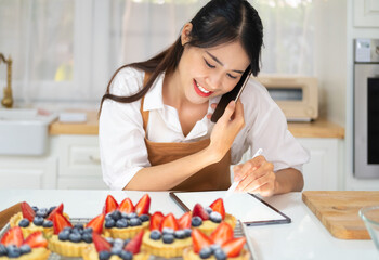 Asian woman bakery shop owner talking on mobile phone with customer about catering bakery order in kitchen. Bakery chef making fruit tart selling online delivery. Small business food and drink concept