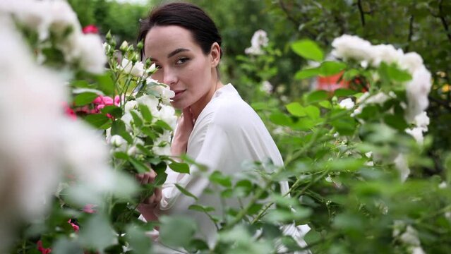 Young woman sitting near bush of white roses in garden and enjoying by it bloom.